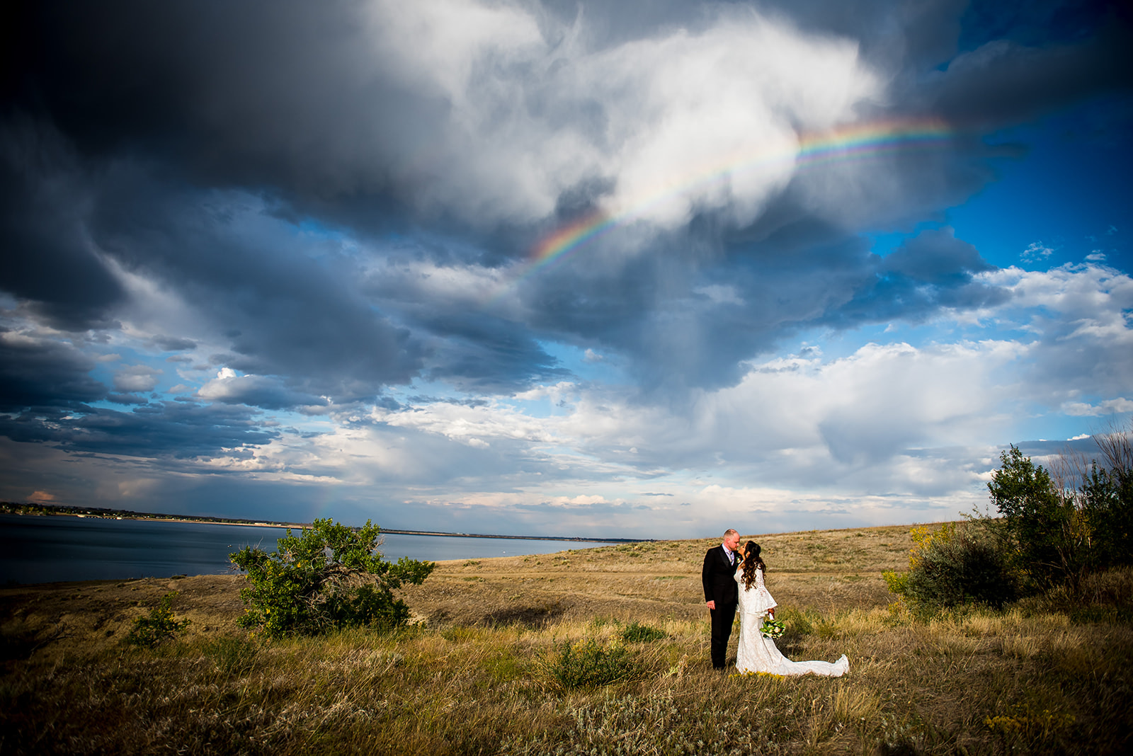 Couple stands facing each other in field with cinematic clouds and a rainbow in the background.