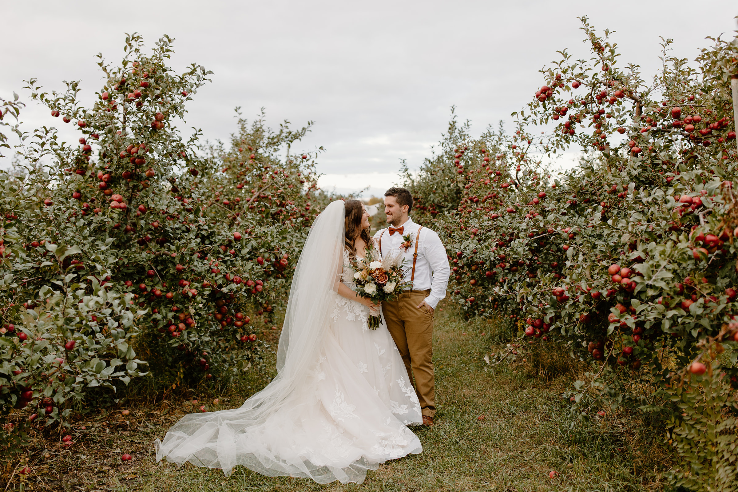 Bride and groom smile at each other in between a row of apple trees