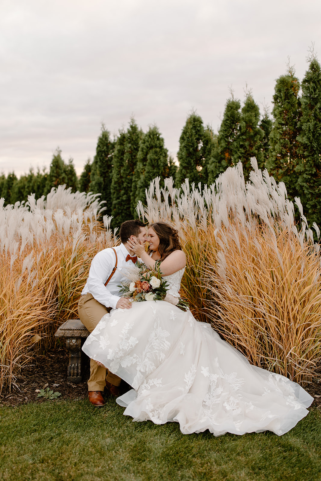 Bride and groom kiss on a bench in front of pampas grass