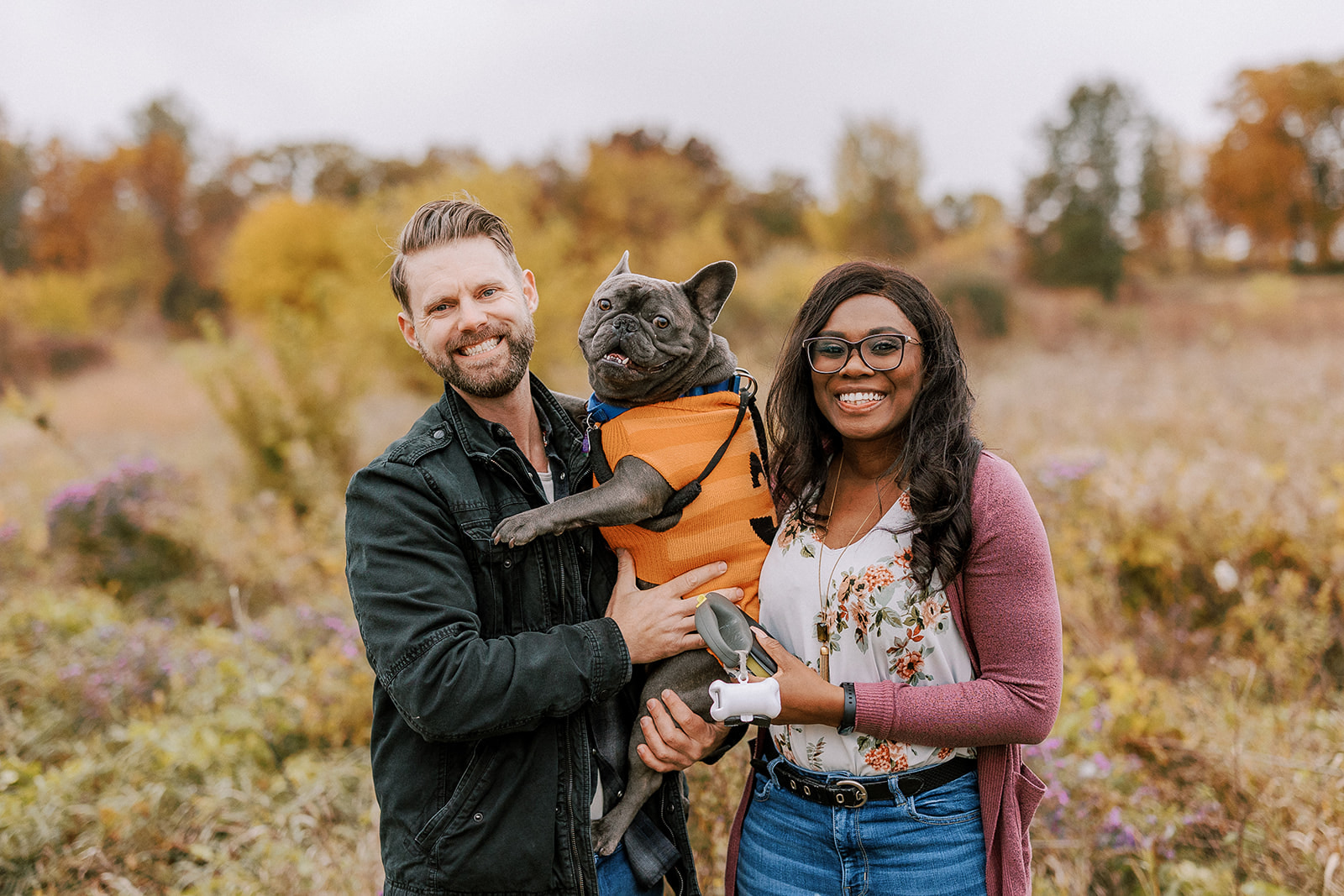 couple smiles at the camera with their french bulldog dressed in a pumpkin costume