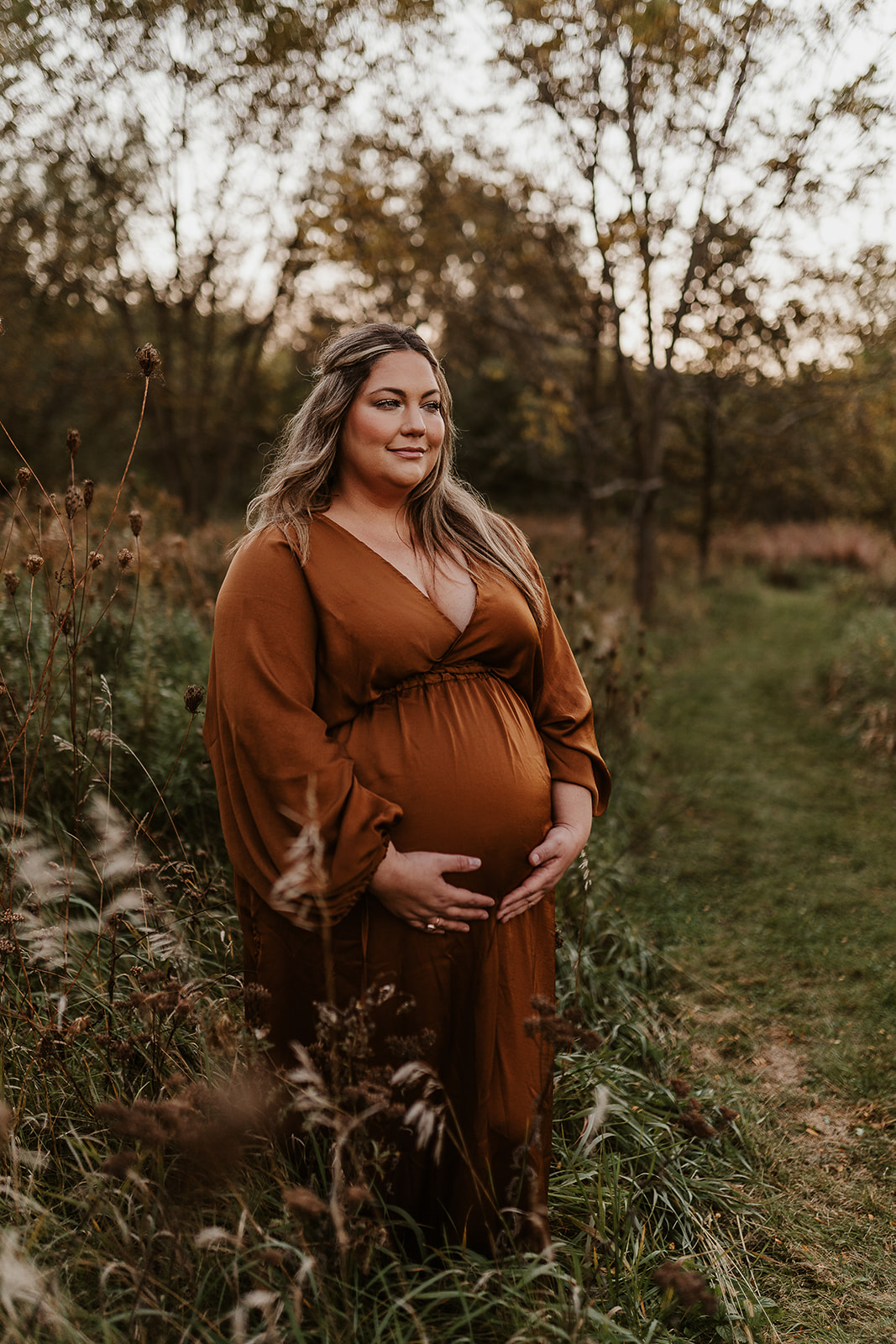 Pregnant mother in a bronze gown holds her belly among the Fall foliage