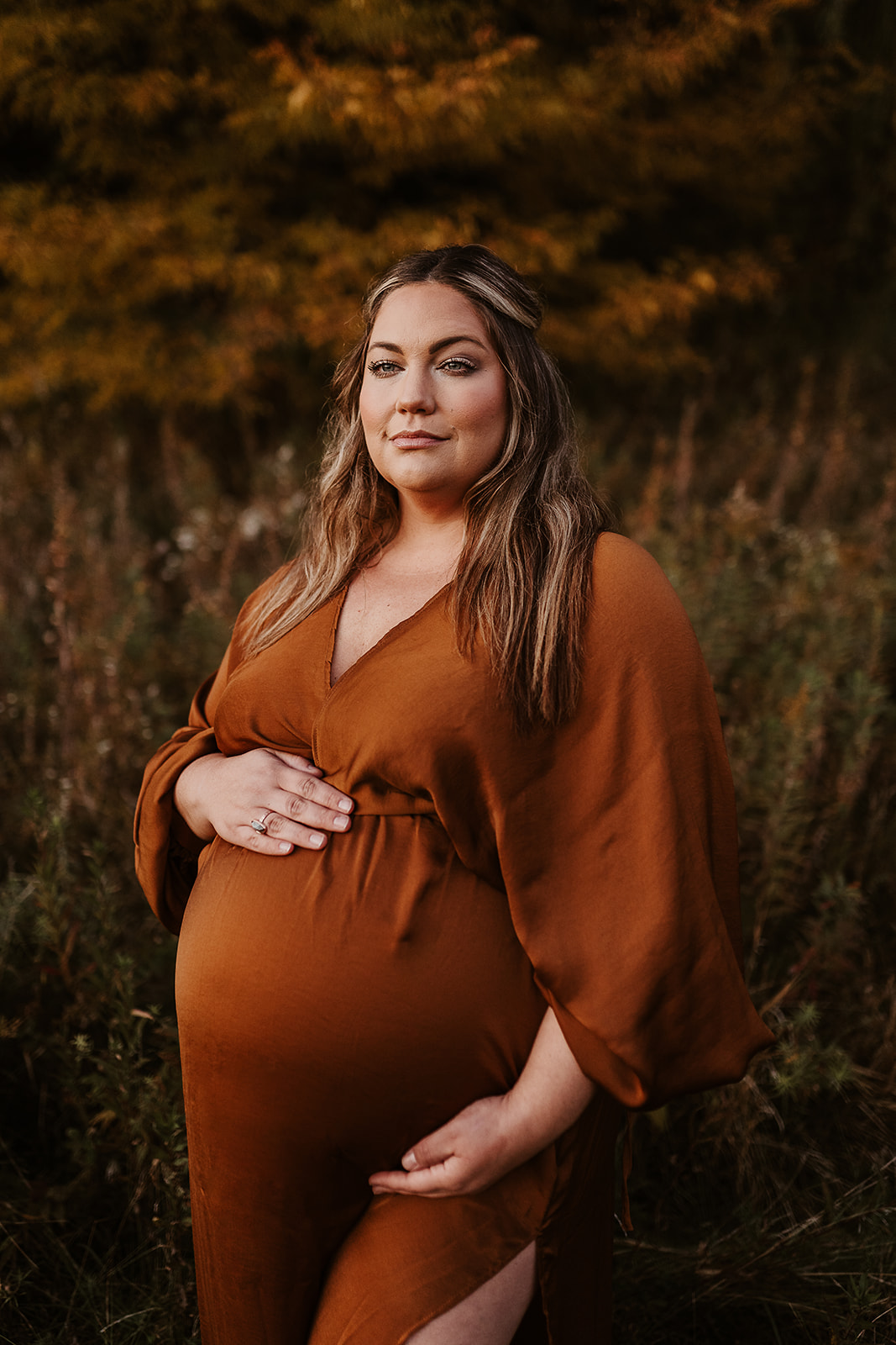 Pregnant mother in a bronze gown holds her belly in front of the golden ochre trees in Autumn