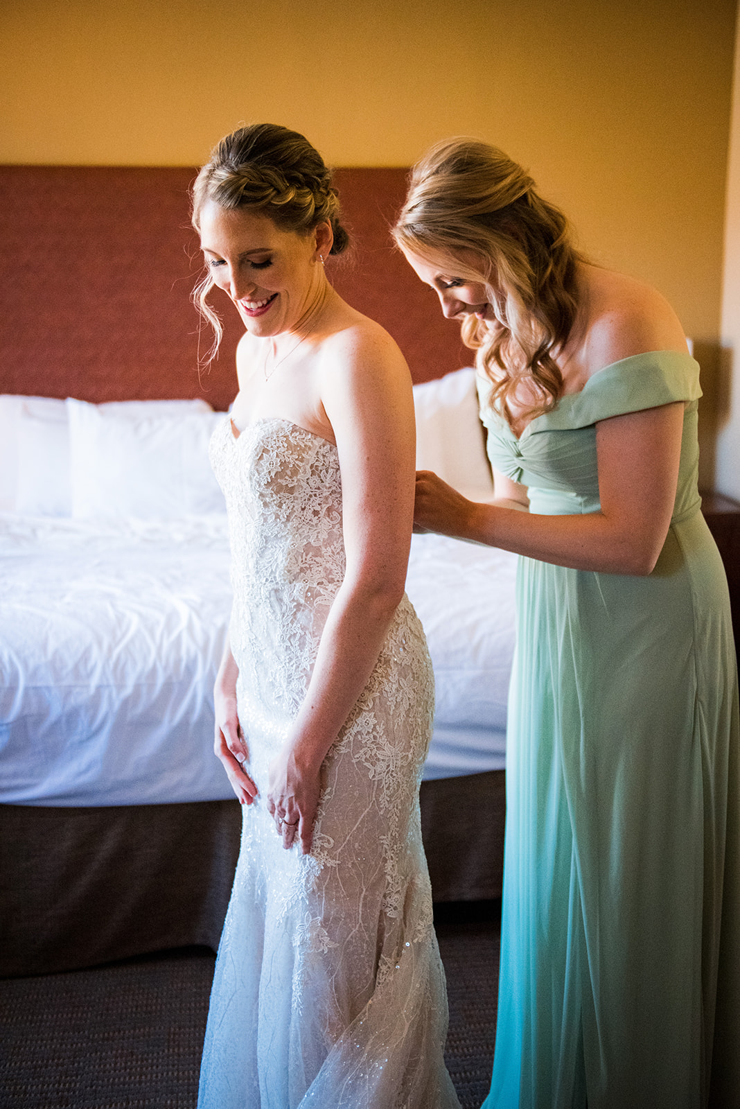 A bridesmaid helps the bride button the back of her dress as the two smile softy.