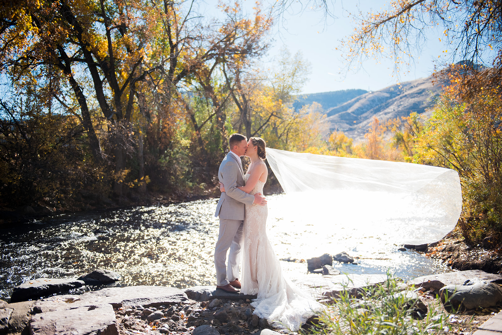 The bride and groom stand kissing on the edge of a beautiful creek with fall leaves in the background.