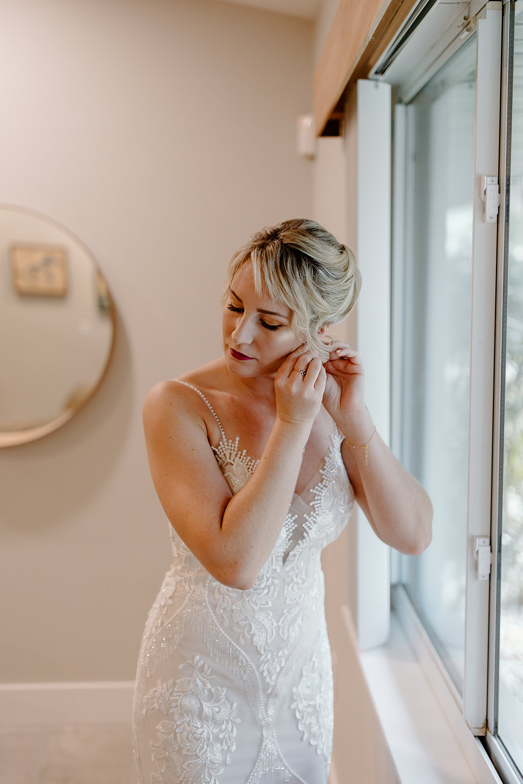 Bride puts her earrings in while standing in front of a window