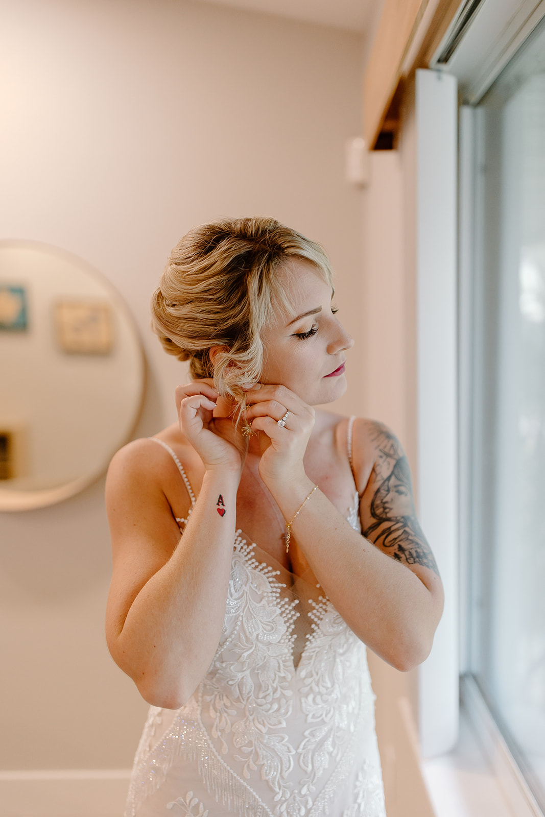 Bride puts her earrings in while standing in front of a window