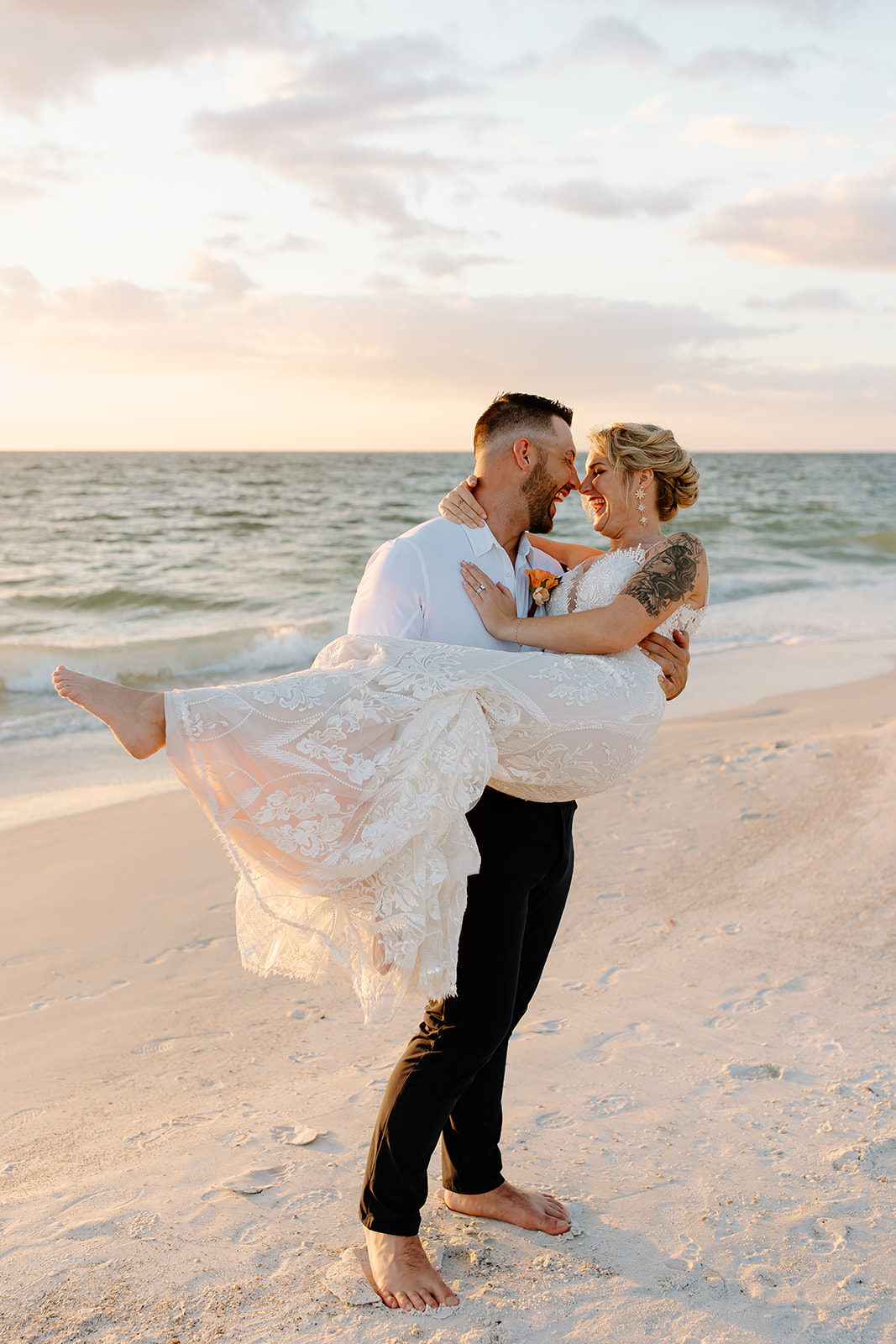 Groom lifts his bride up on the beach in front of the sunset