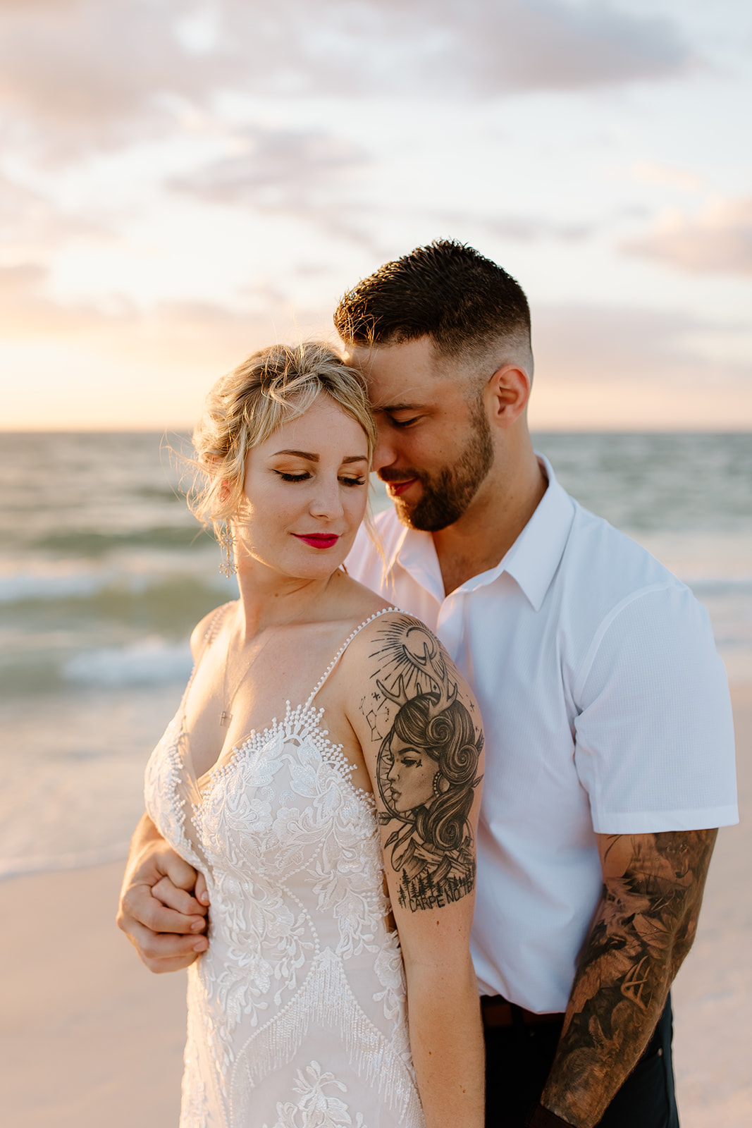 Bride stands in front of her groom on the beach in front of the sunset
