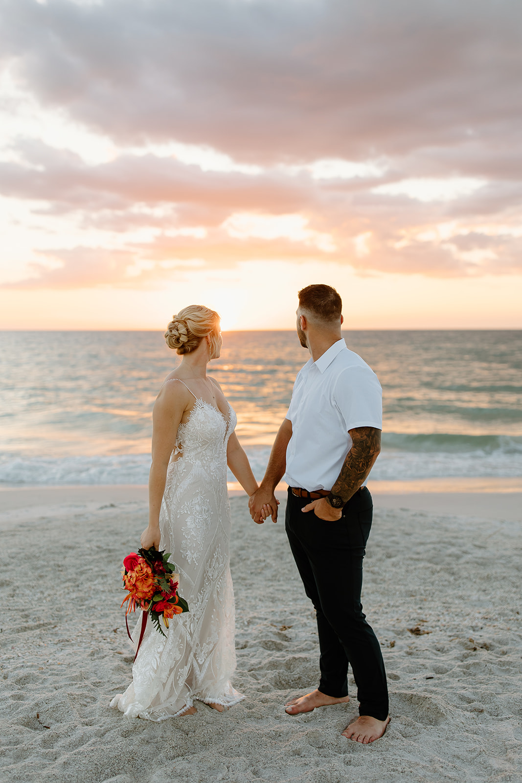 Bride and groom hold hands while walking on the beach in front of the sunset