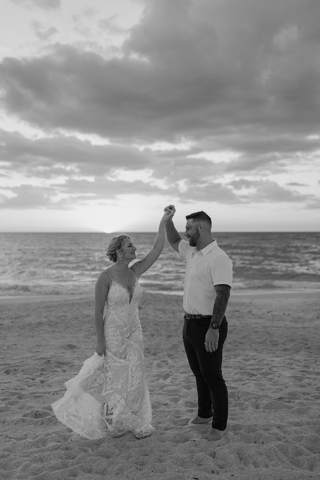 Bride and groom hold hands while walking on the beach in front of the sunset