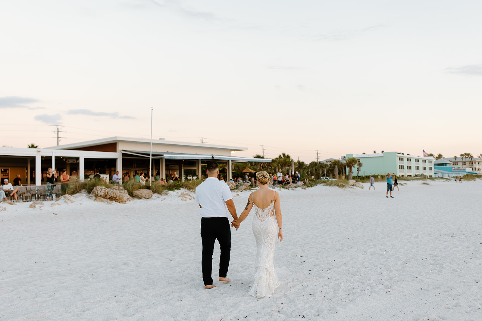 Bride and groom hold hands while walking on the beach towards their guests