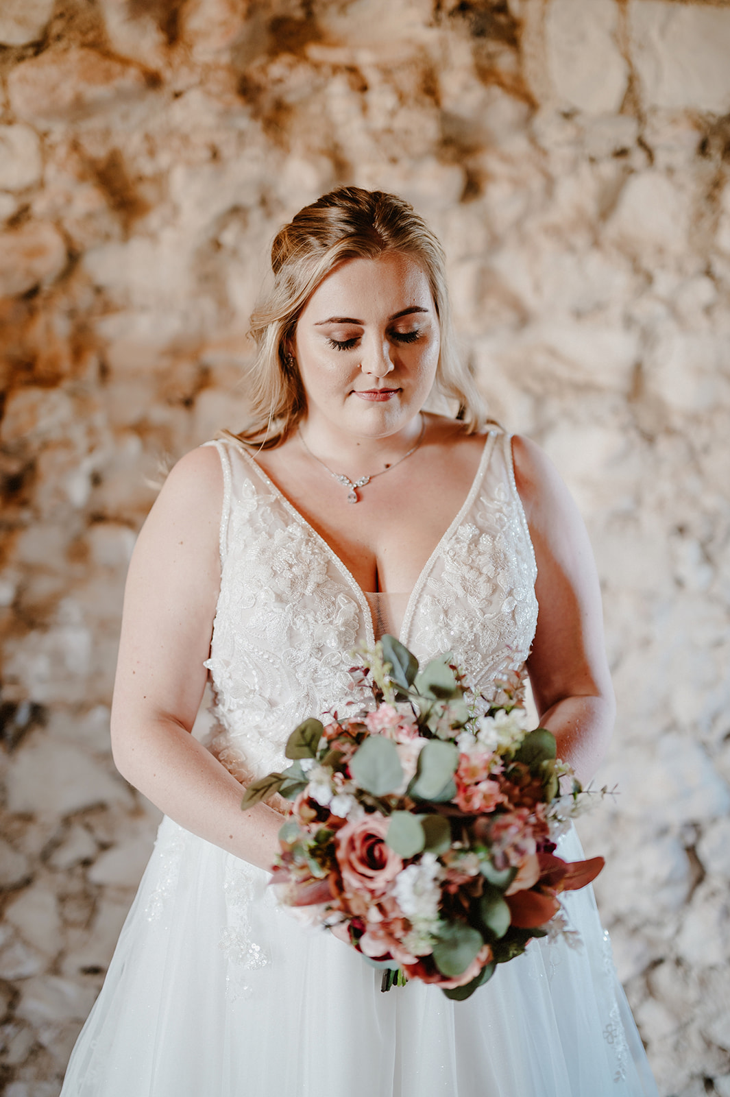 beautiful bride posing for wedding photography looking down at her flowers in front of a rustic brick wall at pentney abbey in natural light