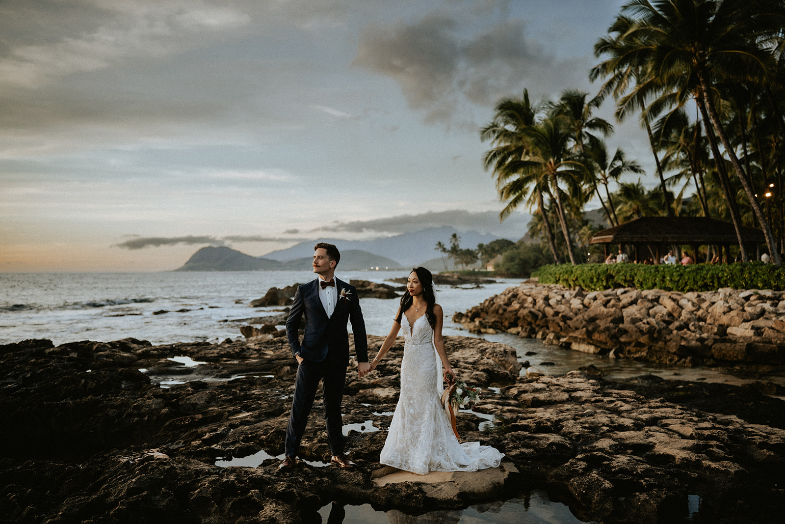 Bride and groom on the rocky shore of the beach fronting Lanikuhonua during sunset