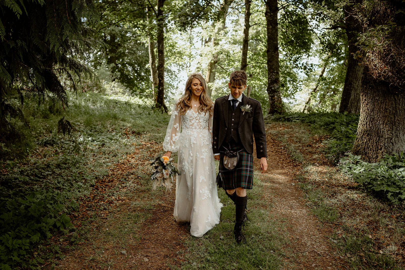 boho bride and groom walk holding hands through a sunny woodland