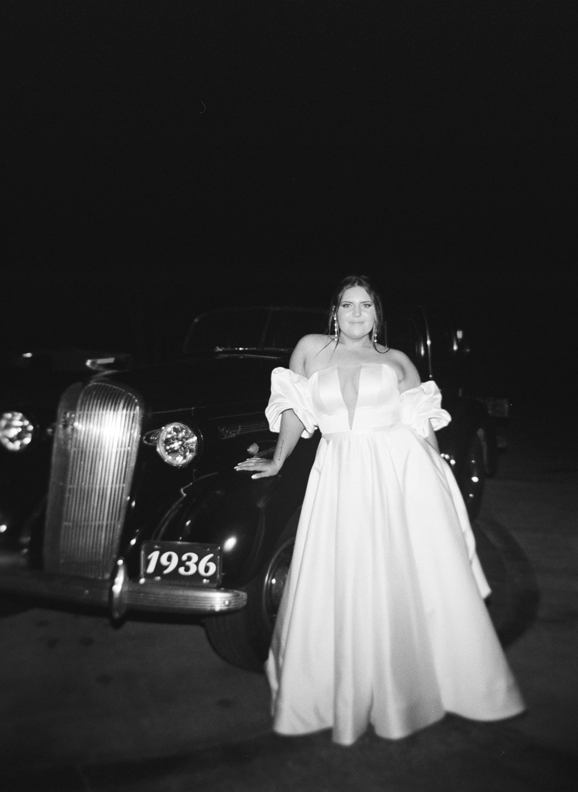 a black and white film photo of the bride in front of her vintage getaway car.
