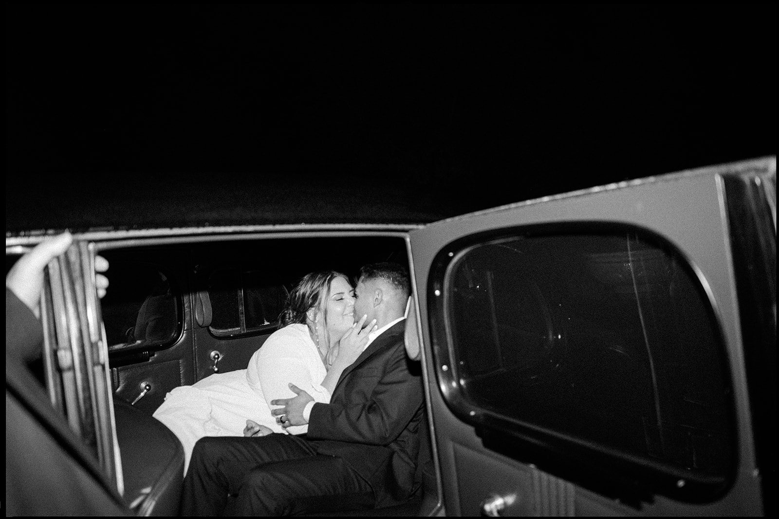 candid black and white photo of the bride and groom sharing a kiss in the back of their vintage getaway car