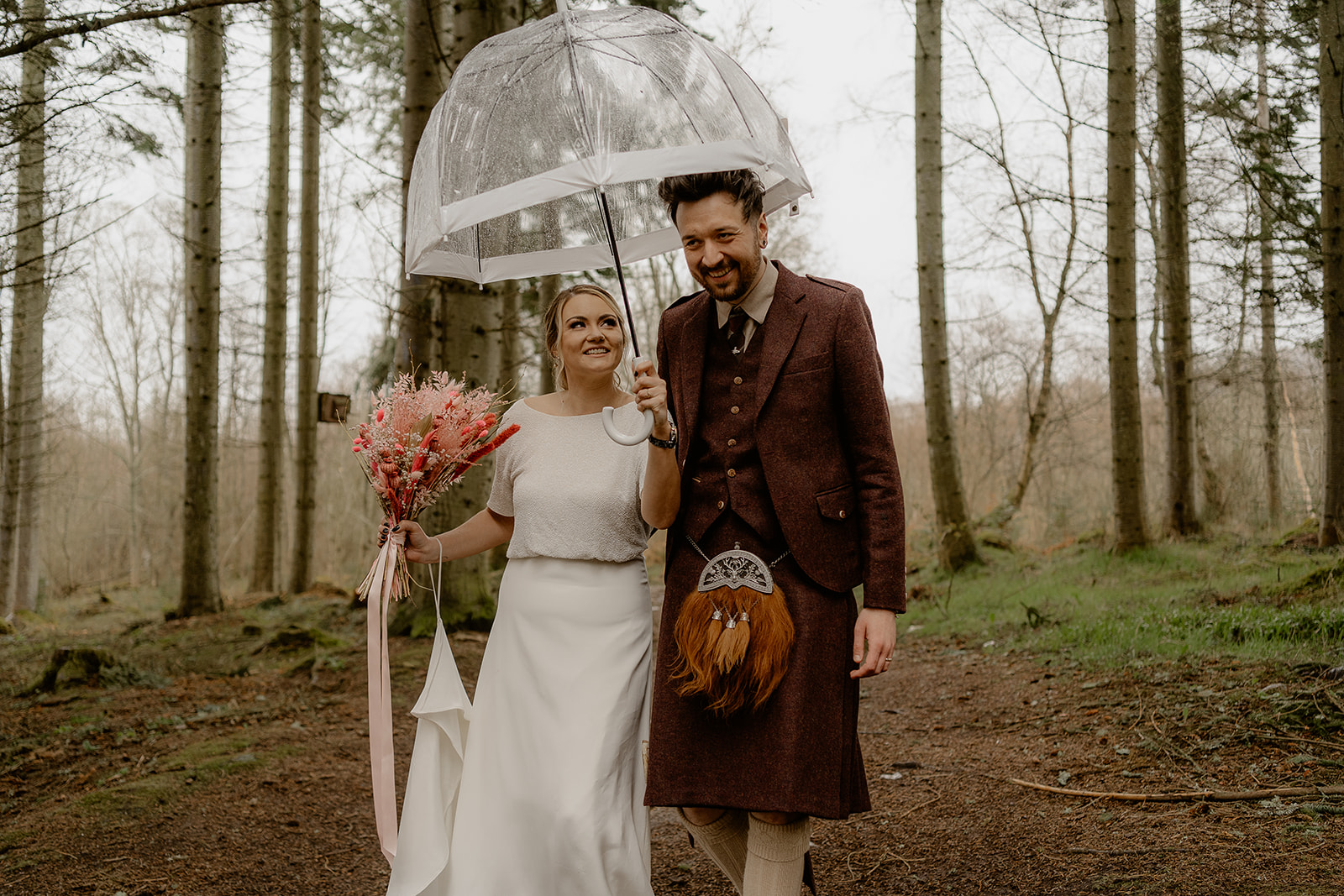 bride and groom walking through woodland underneath an umbrella