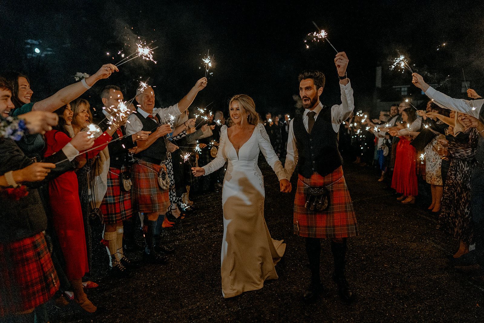 Bride and Groom walking holding sparklers as their guests line up either side and cheer