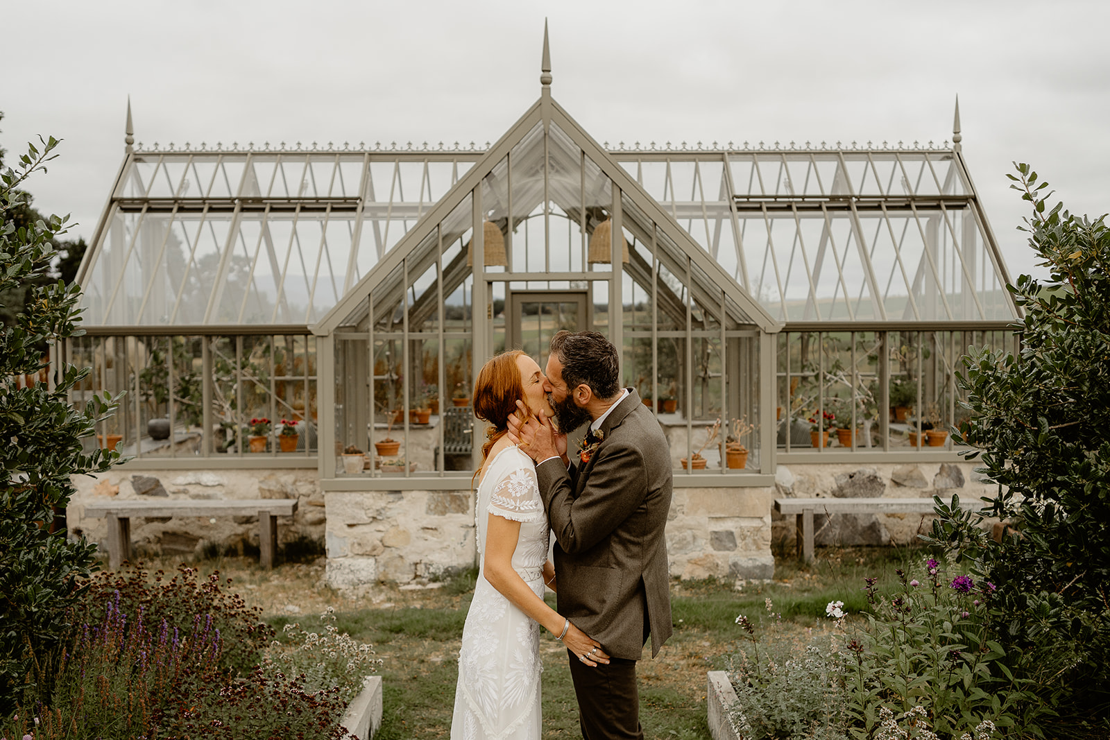 bride and groom kiss in front of victorian greenhouse surrounded by foliage