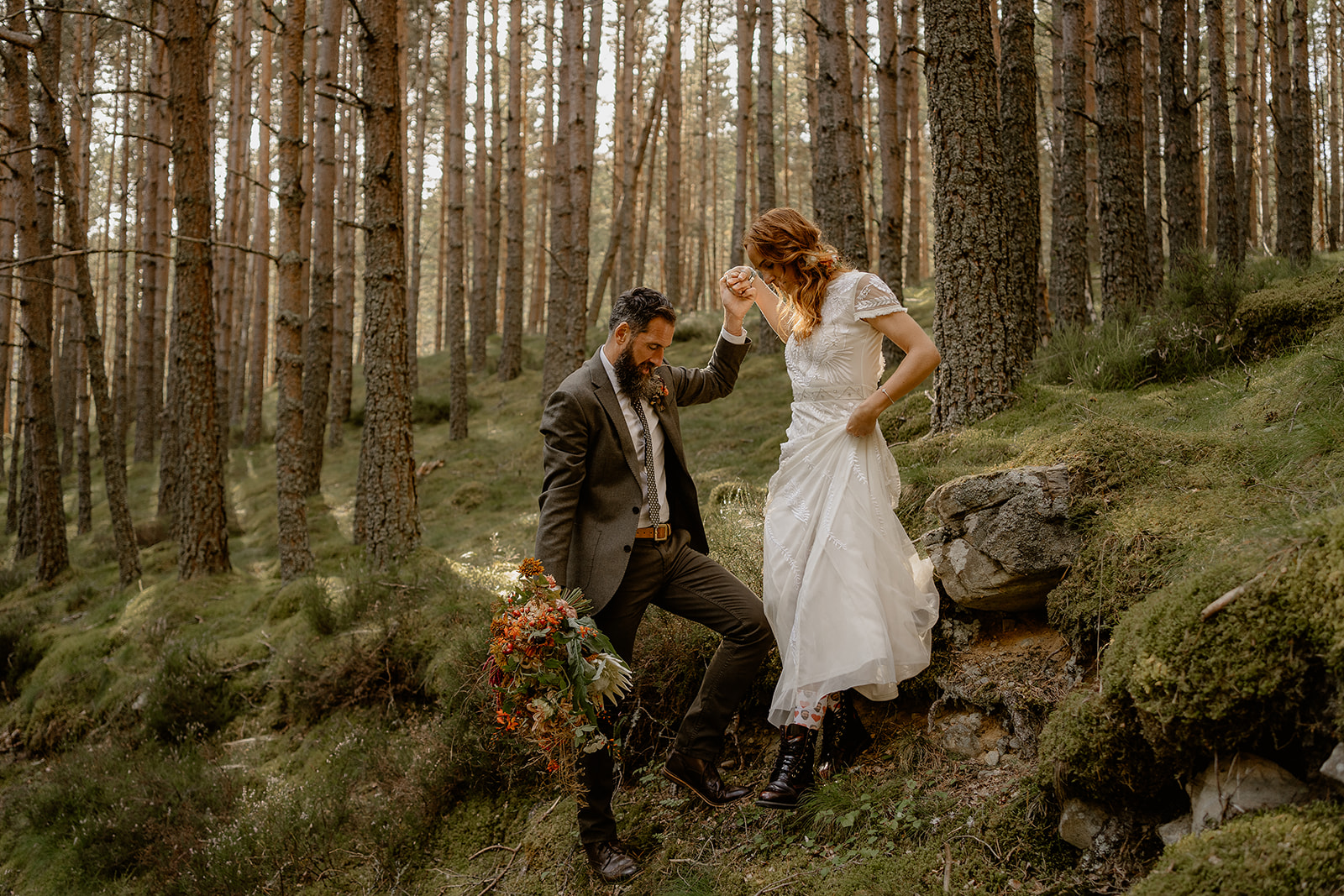 groom helps bride down steep mossy bank in woodland by holding her hand. He holds her bouquet of flowers while she walks