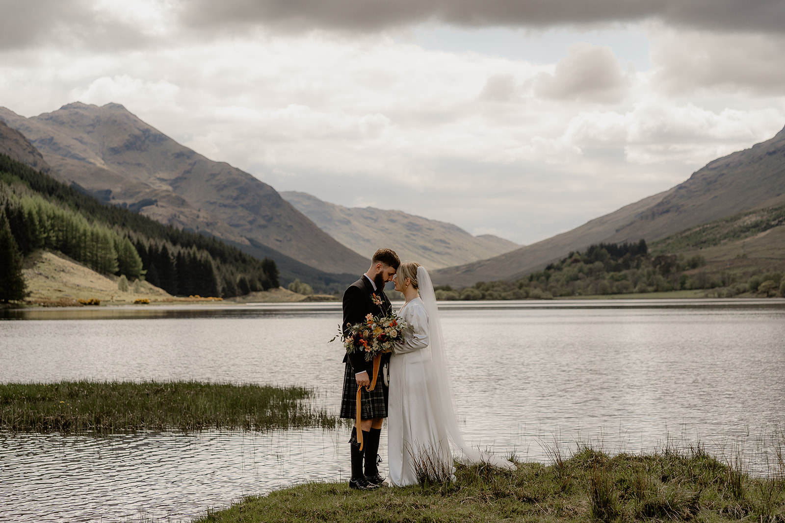 highland elopement at monachyle mhor. couple embrace next to loch with dramatic mountains and sky behind them