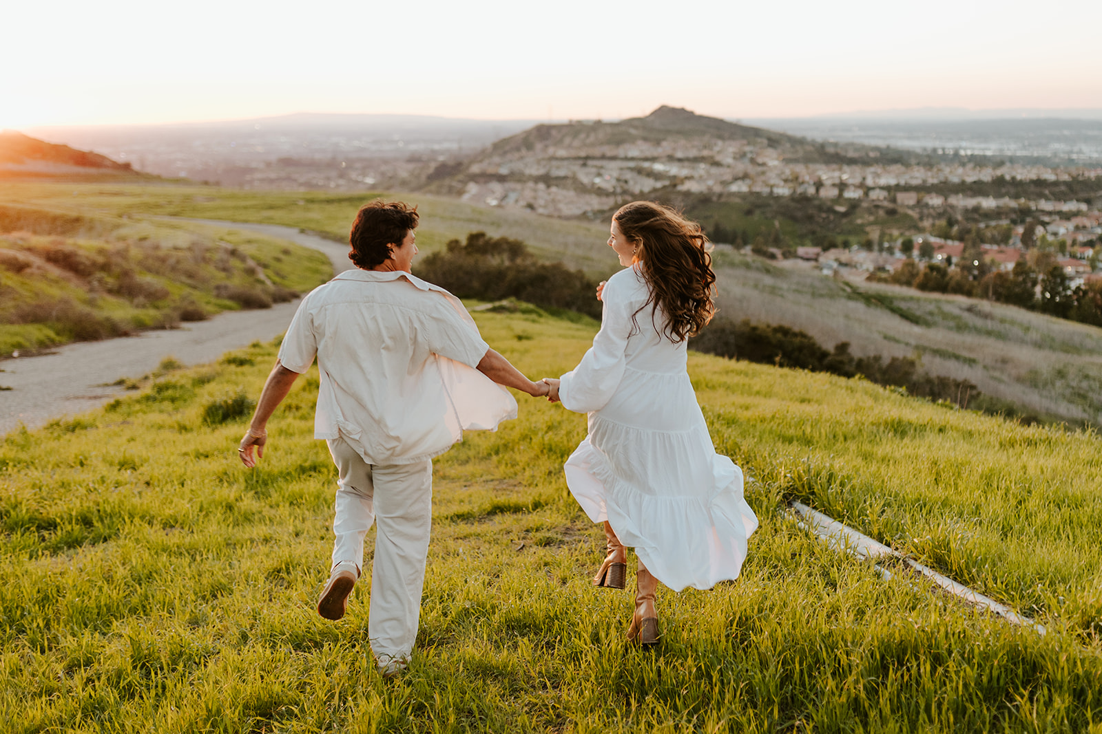 A couple in linen clothes runs together at sunset through the California hills.