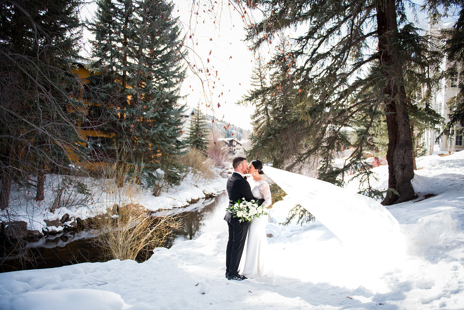 Bride and groom standing in the snow kissing as the bride's veil blows in the wind.