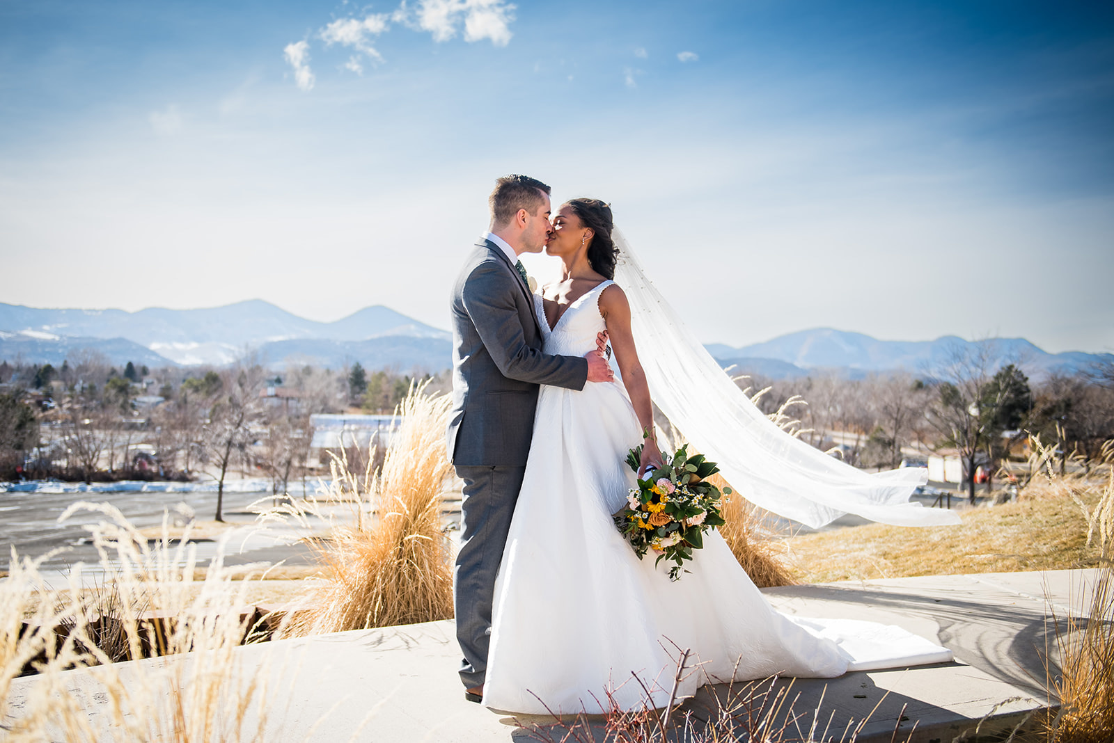 Bride and groom share a kiss with a mountain backdrop as the veil floats in the wind.