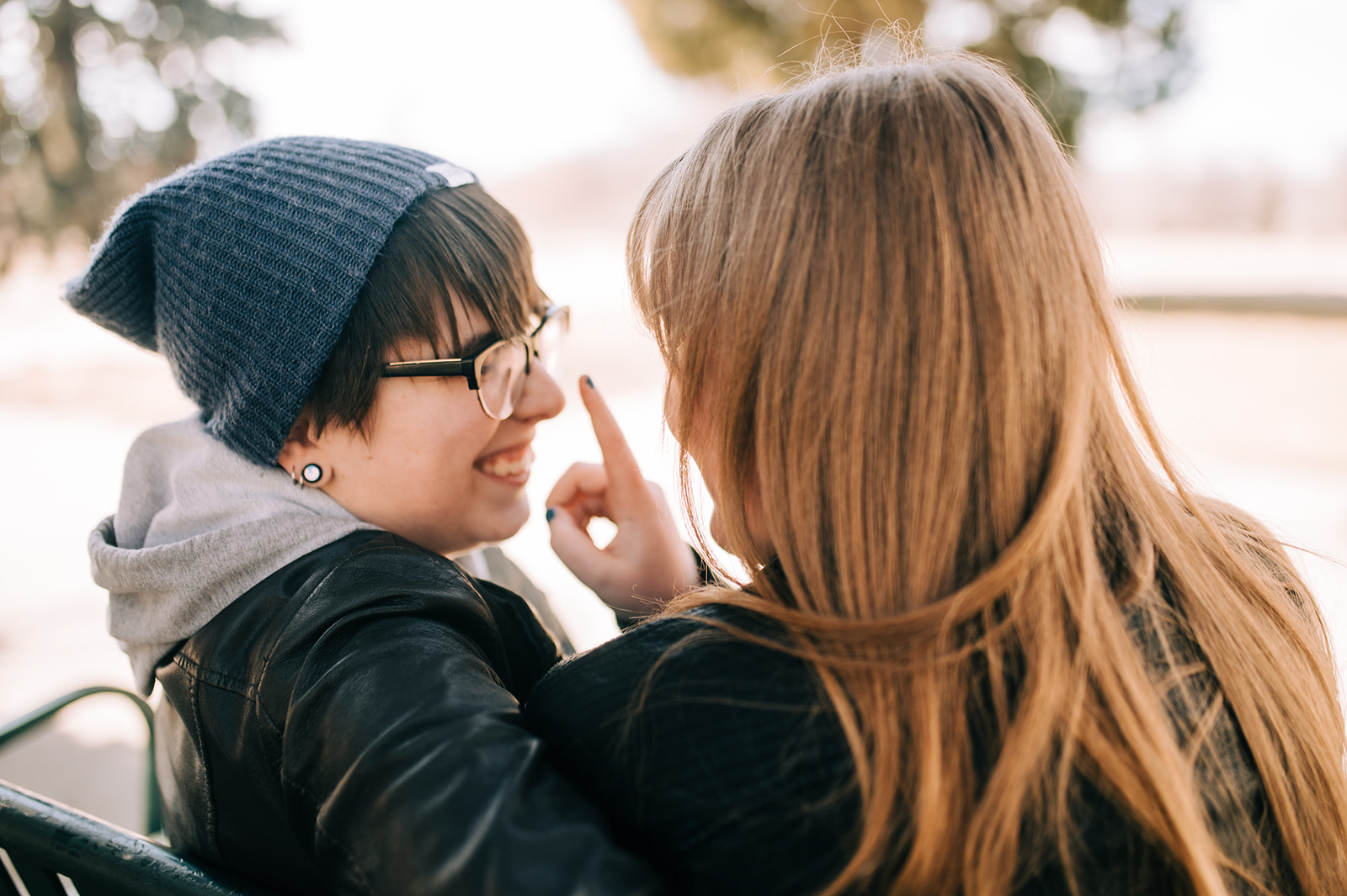 photo from behind of two women sitting on a park bench while one boops the other's nose