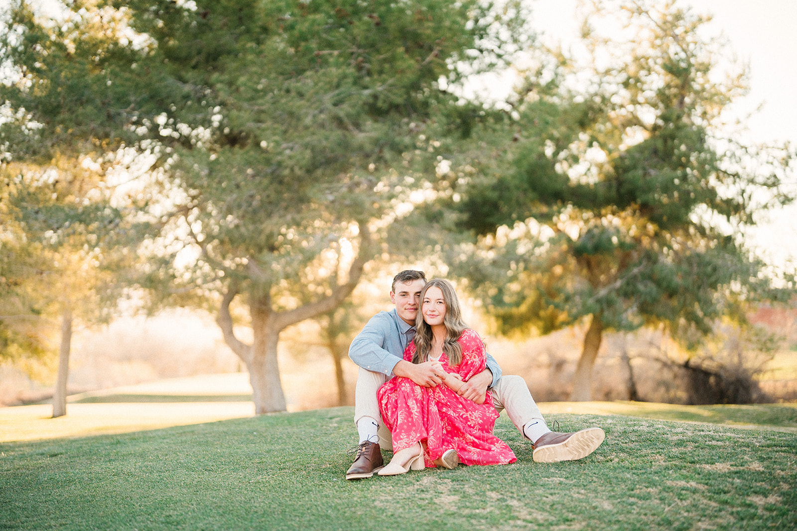 engaged couple sits on grass with trees