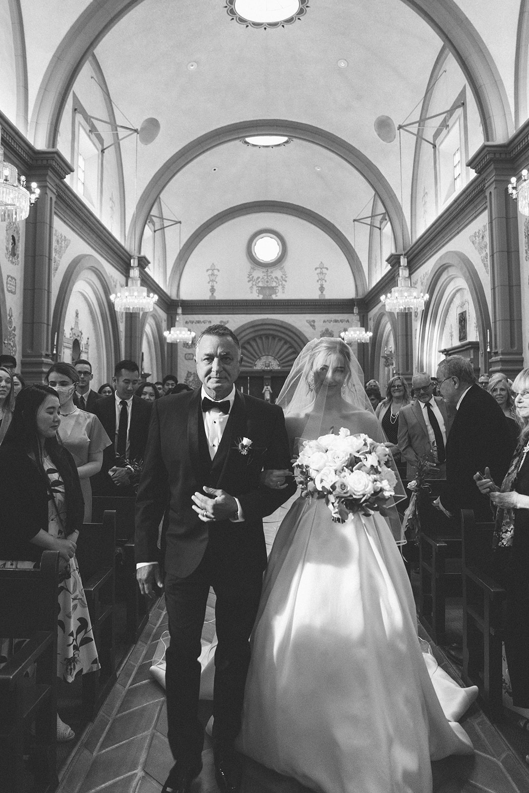 Bride walks down the aisle with her dad during her wedding ceremony at the Mission Basilica in San Juan Capistrano, CA. 