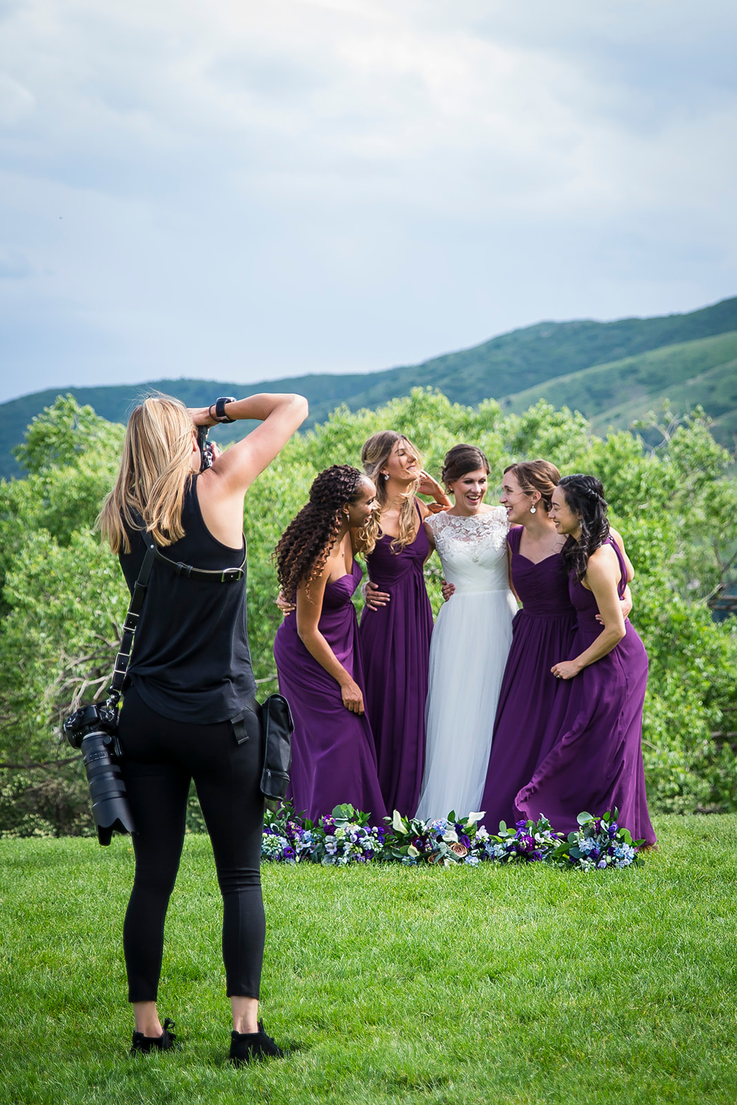Wedding photographer captures group of bridesmaids and bride posing for photo in green, grassy field.
