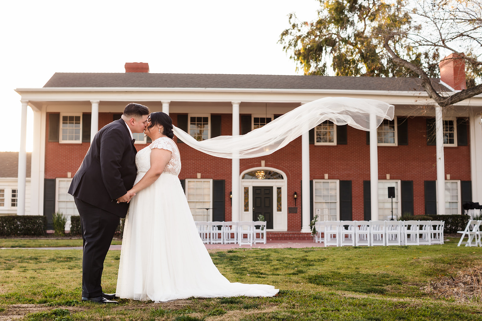Bride and groom share a kiss in front of Crestmore Manor as bride's veil flows in the wind