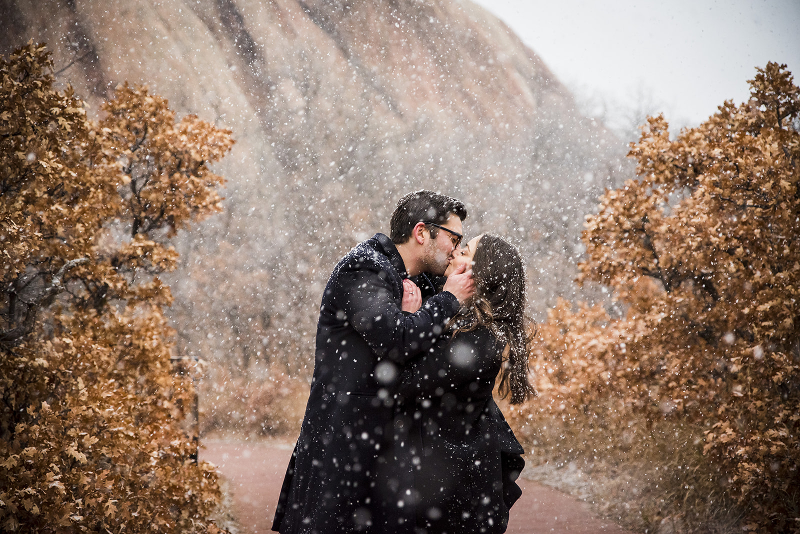 Couple kisses each other in the snow with red rock mountain formation behind them.