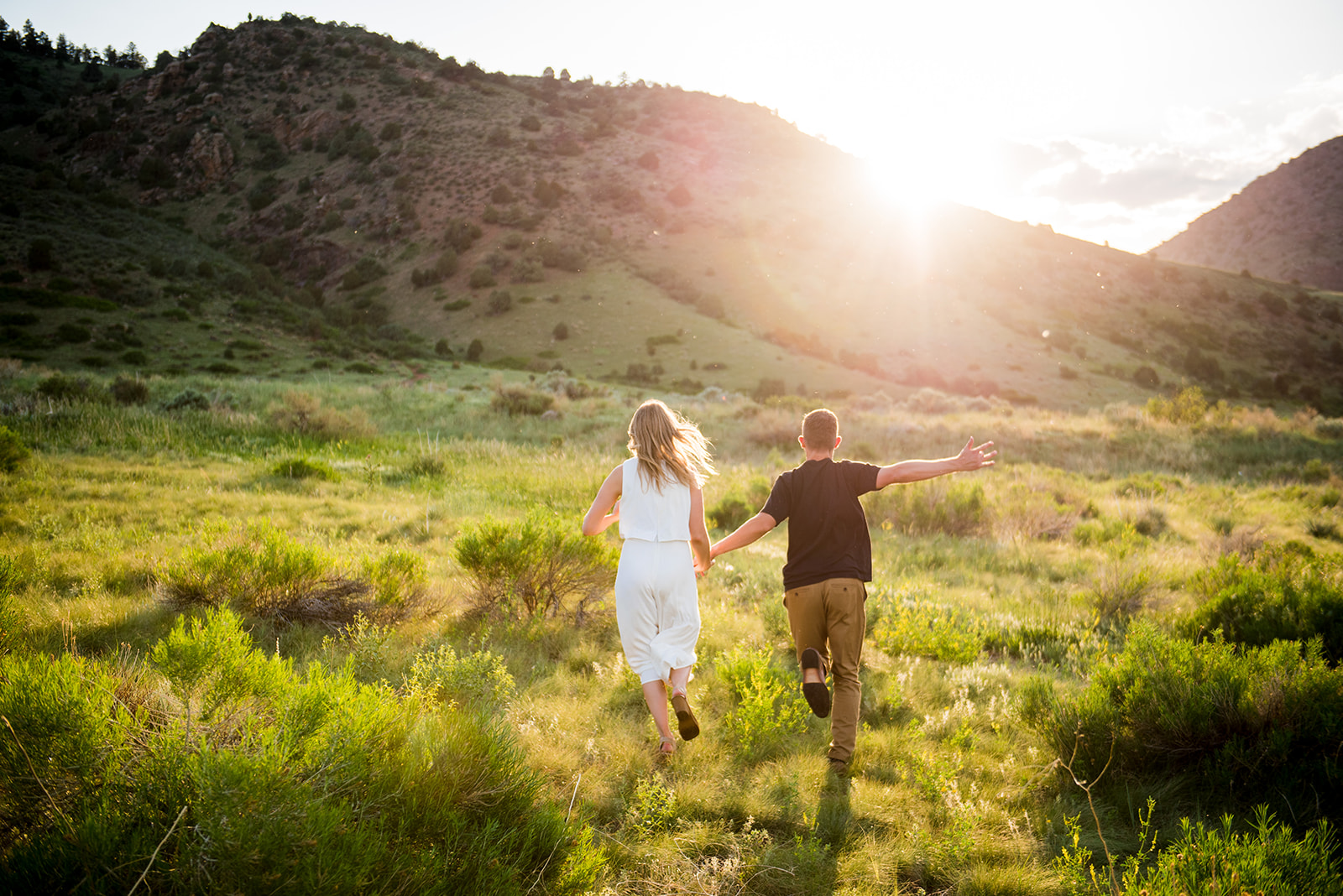 Couple runs through green, grassy field with backs facing camera and sunset going behind a mountain in front of them.