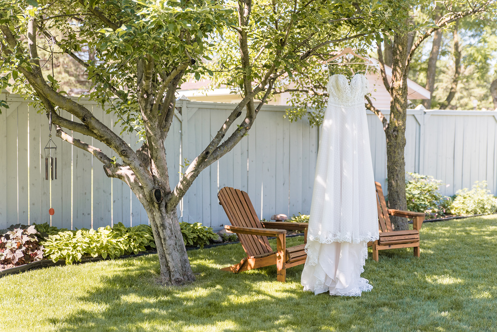 A wedding dress hangs from a tree with spring leaves and two adirondack chairs in the background