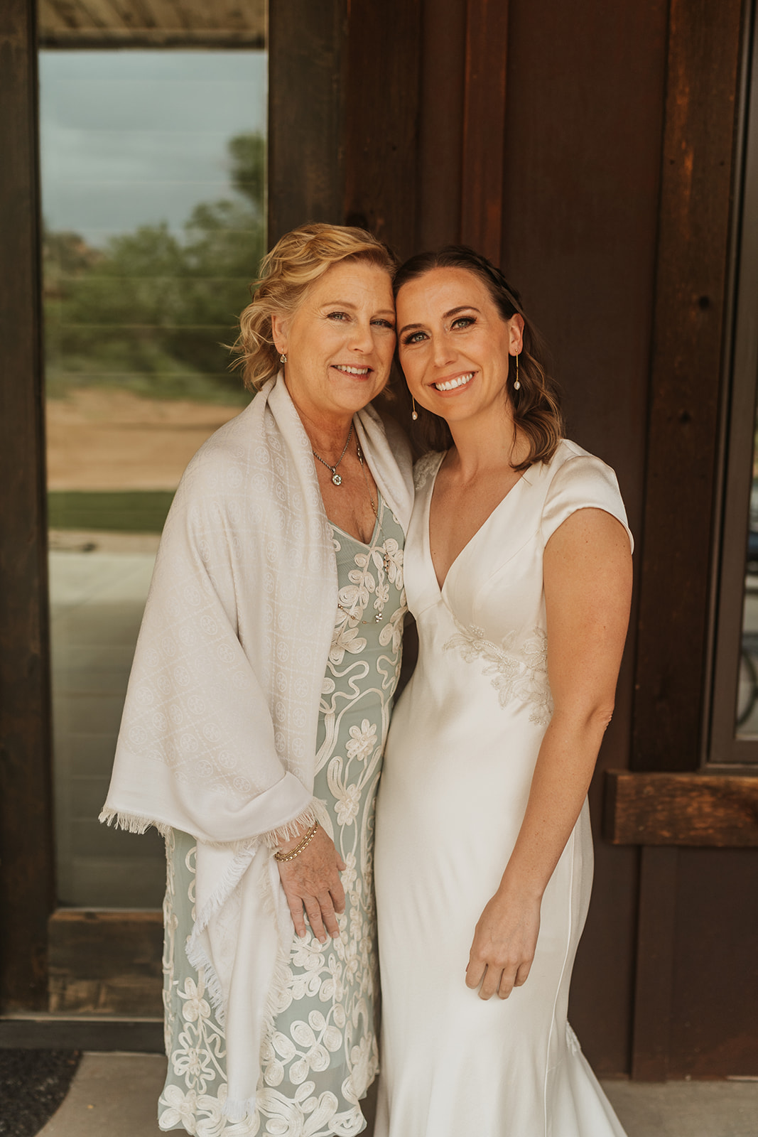 Mother her daughter, the bride, standing next to one another with a wooden wall behind them 