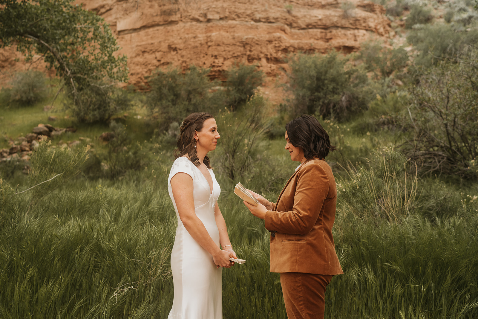 Two brides standing facing one another and reading vows to each other 