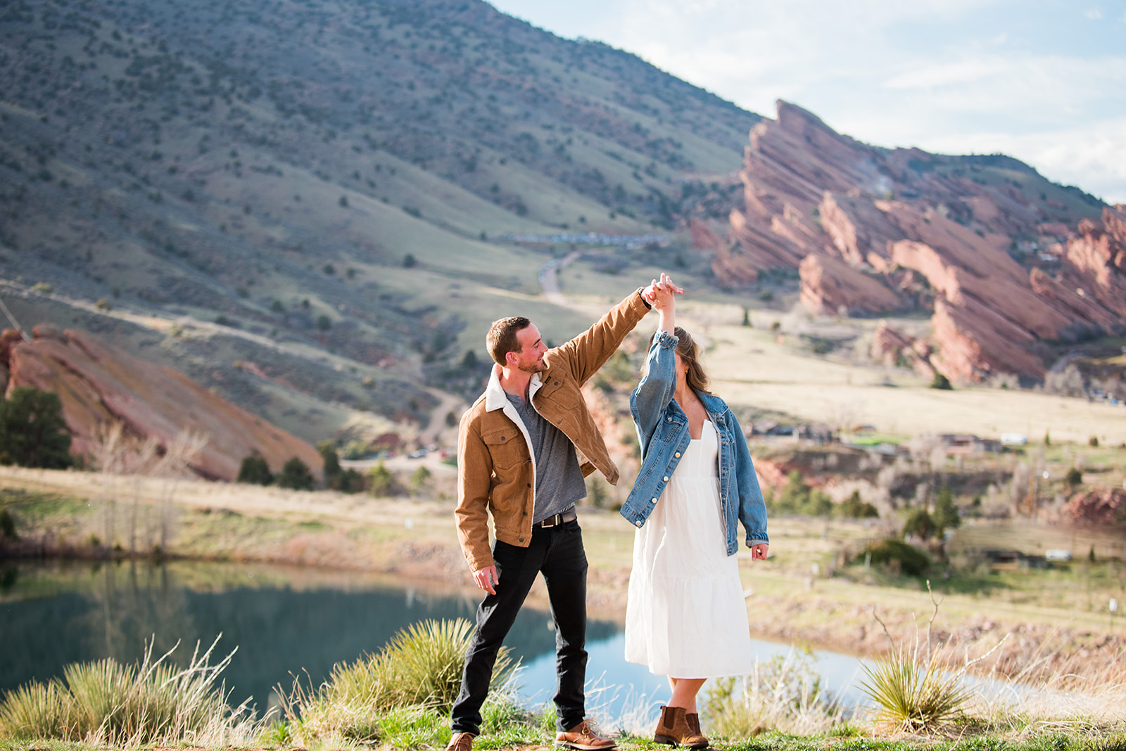 Man twirls his fiancée in front of a lake and red rocks at Mount Falcon Colrado.