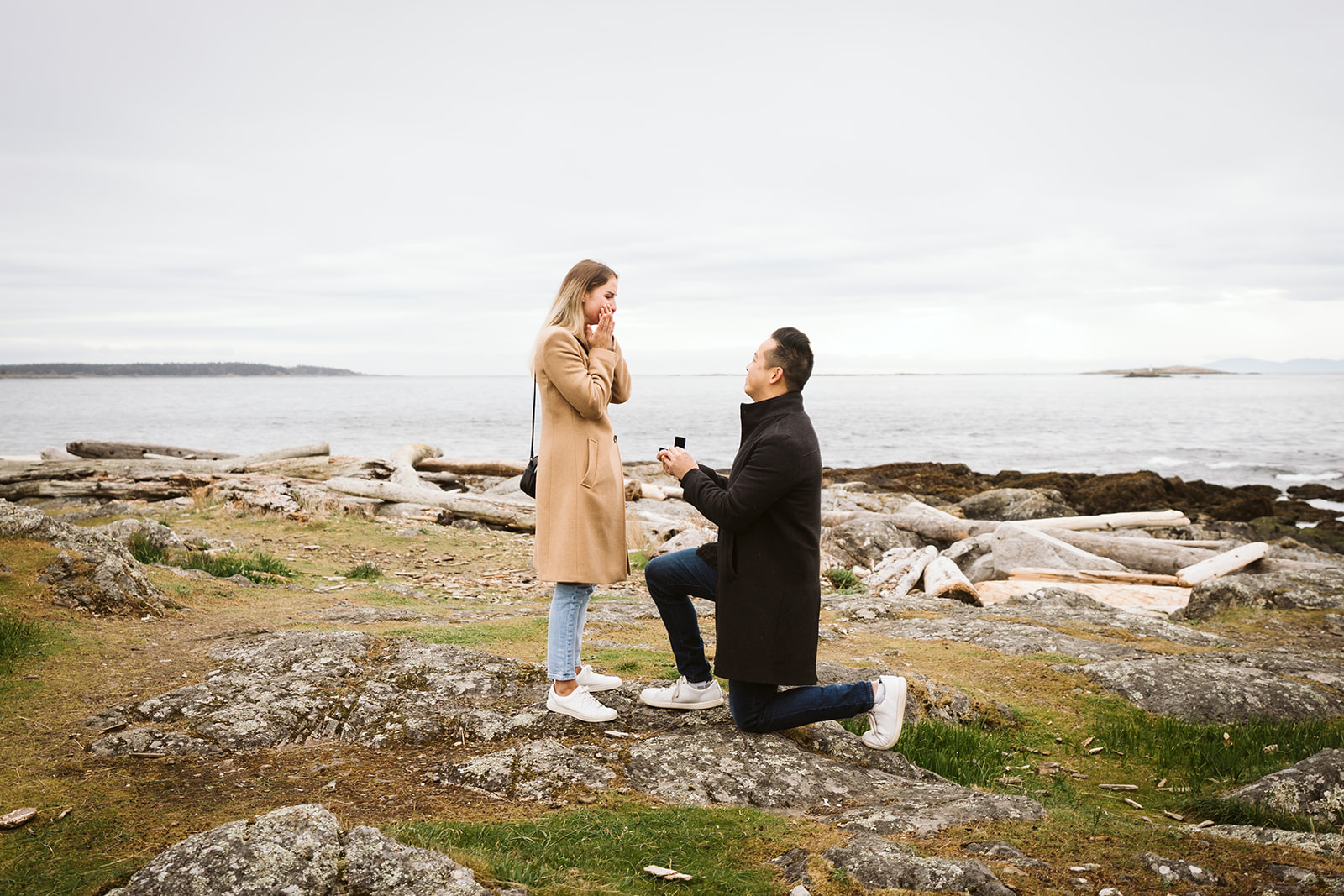 Man kneeling on rocks as he proposes to his girlfriend. She has her hands up, covering her face.