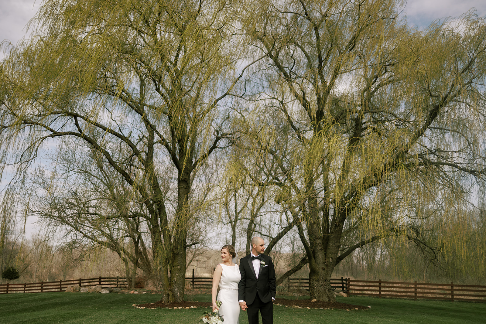 bride and groom stand in front of willow tree at harvest moon pond on their wedding day.